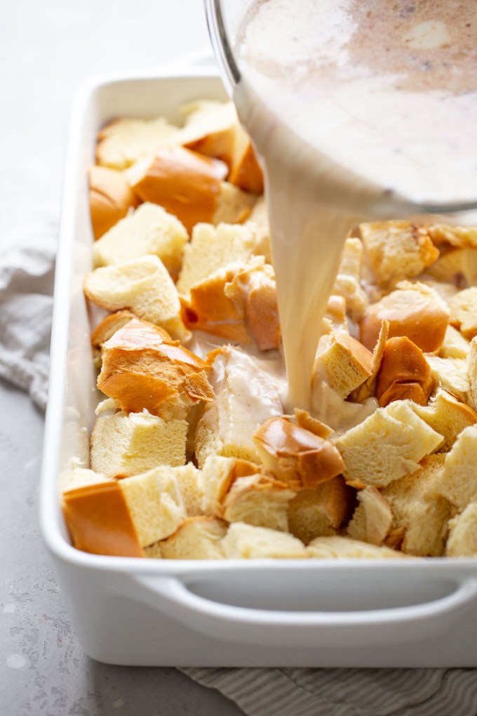 The egg custard mixture being poured over cubed bread in a 9x13 pan.