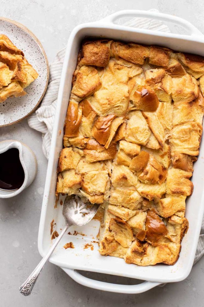 An overhead view of bread pudding in a 9x13 pan. A slice of bread pudding and small container of syrup rest beside it.