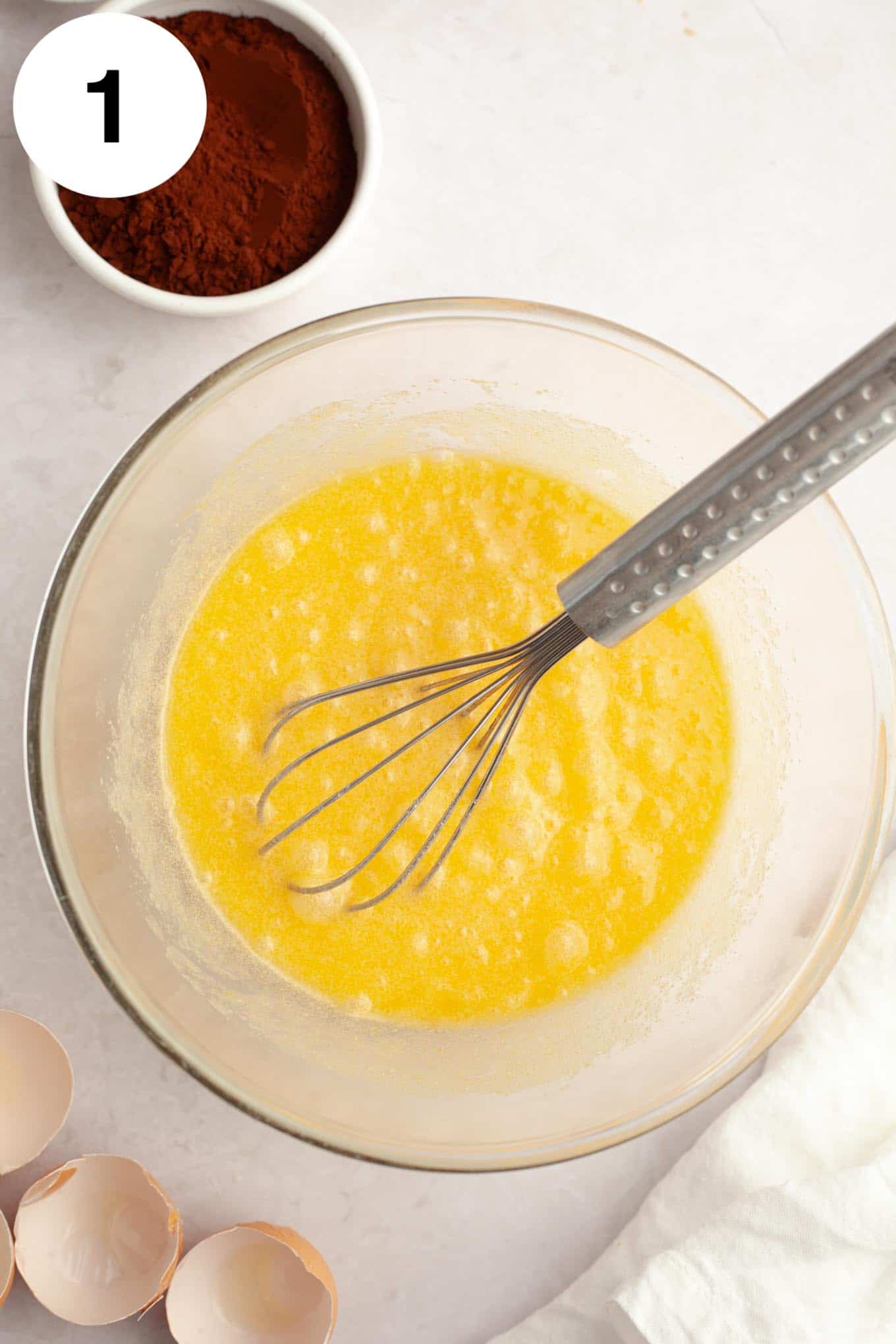 An overhead view of melted butter and sugar in a glass mixing bowl, with a whisk. 