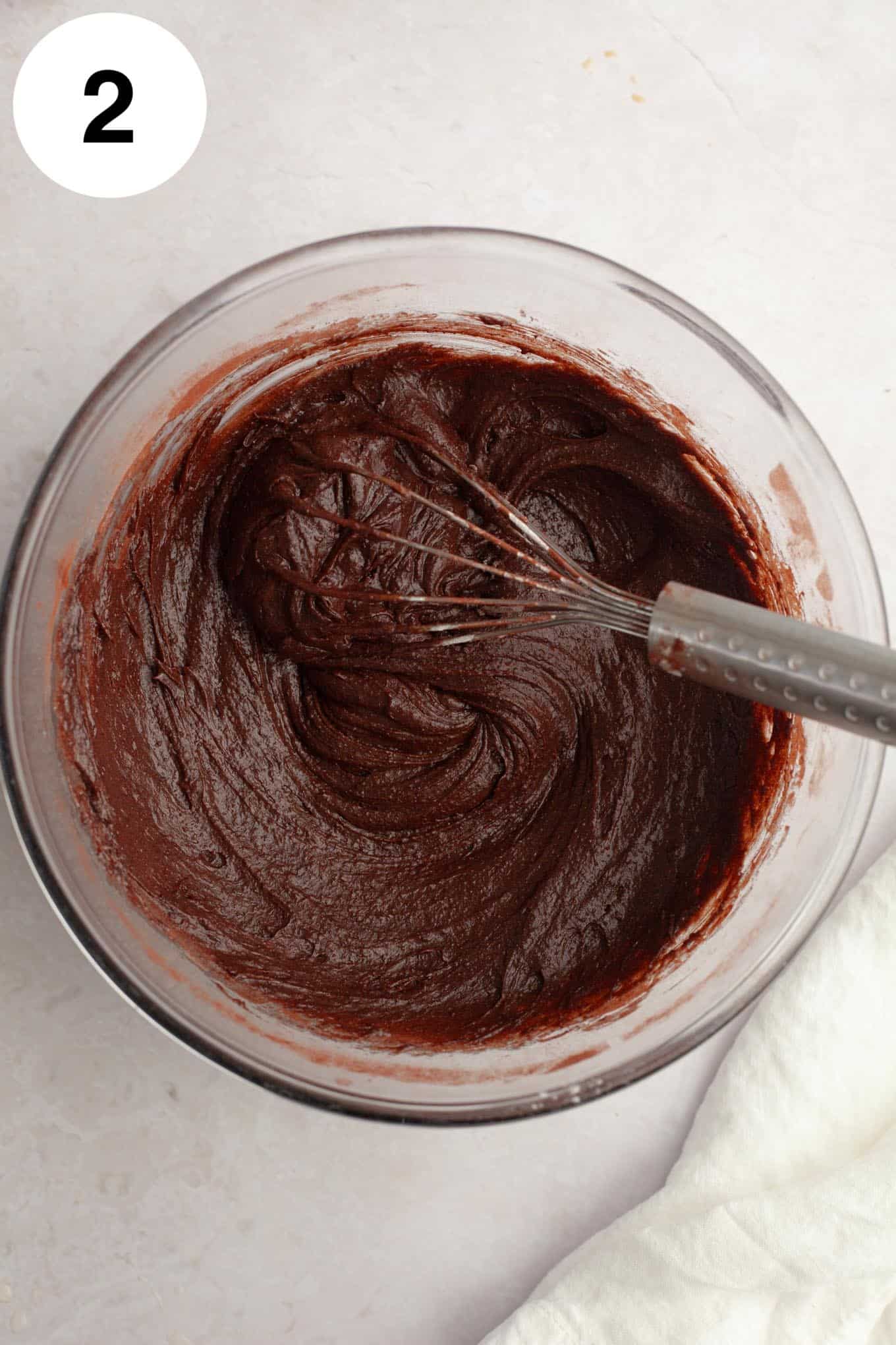 An overhead view of brownie batter in a glass mixing bowl, with a whisk. 