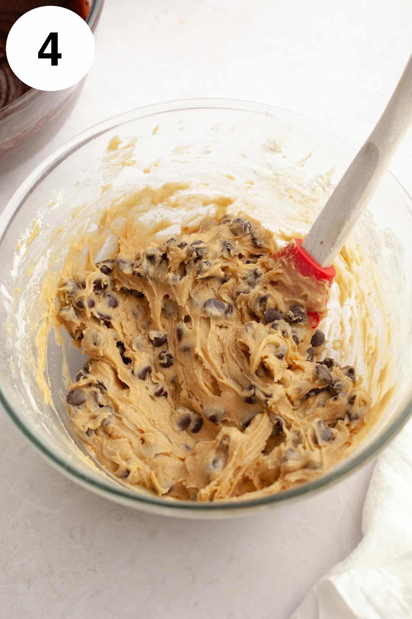 An overhead view of chocolate chip cookie dough in a glass mixing bowl, with a whisk. 