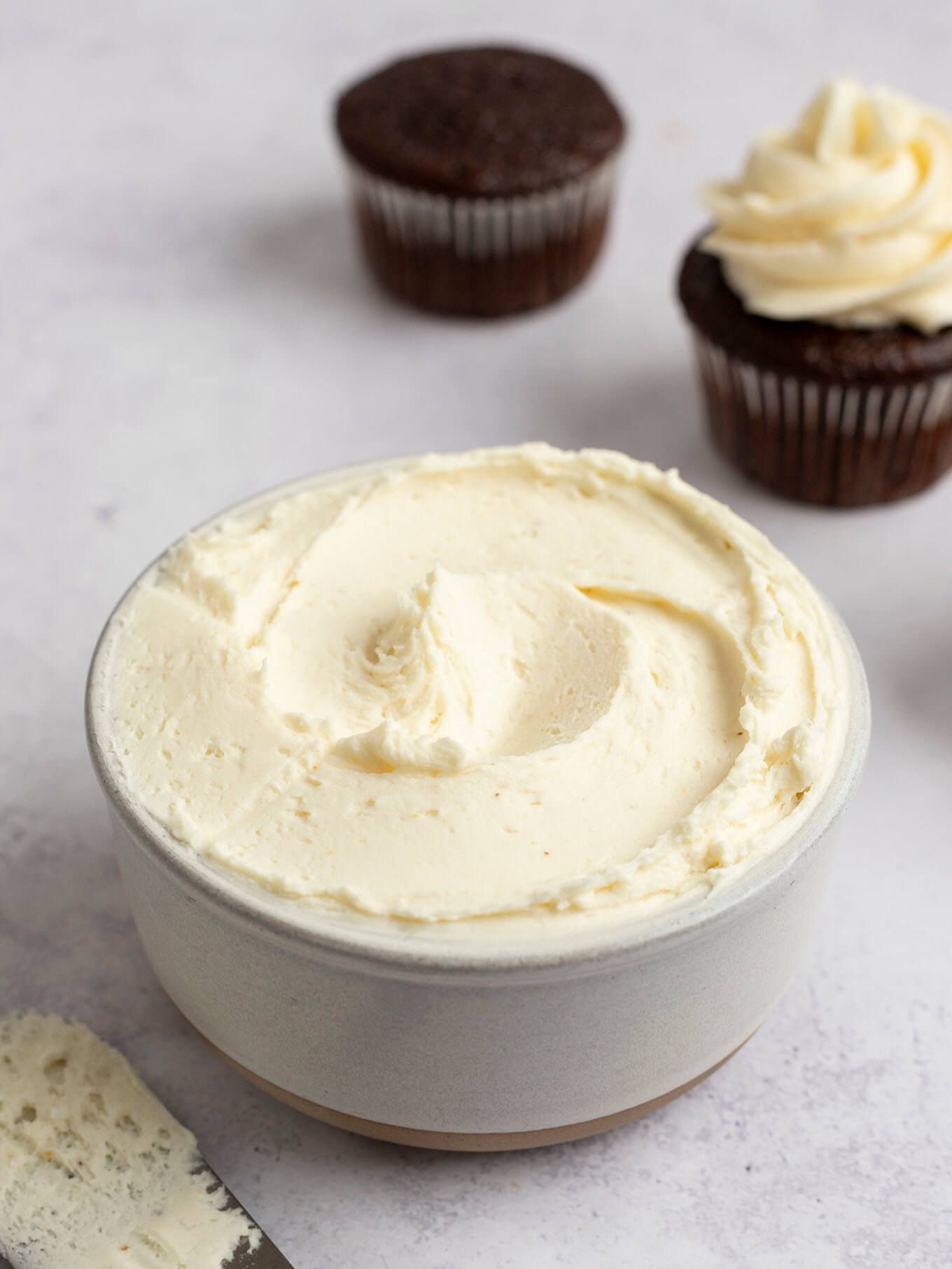 Brown butter frosting in a white bowl. Two chocolate cupcakes are sitting near the bowl.