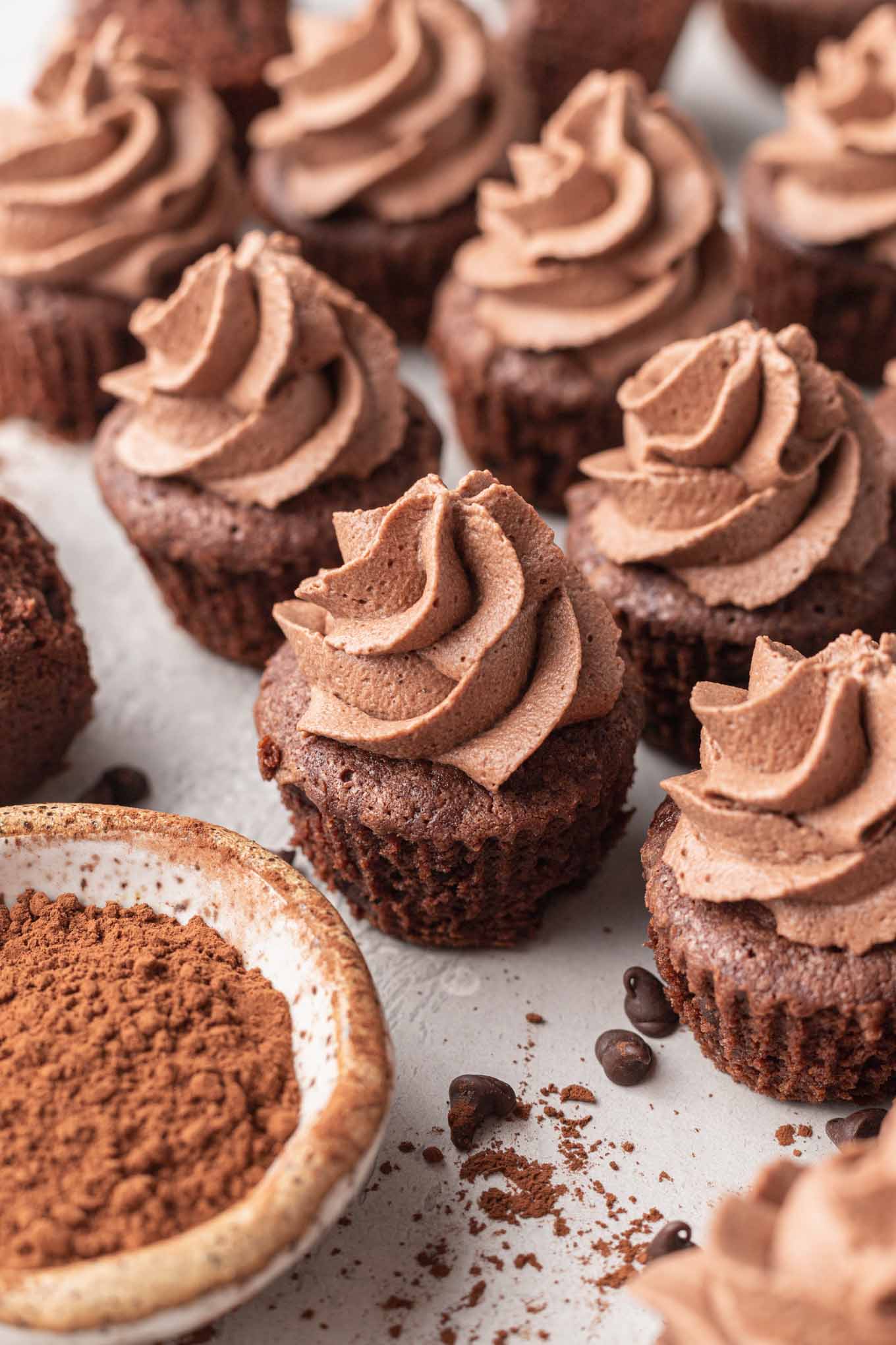 Mini brownies topped with chocolate whipped cream, next to a bowl of cocoa powder. 