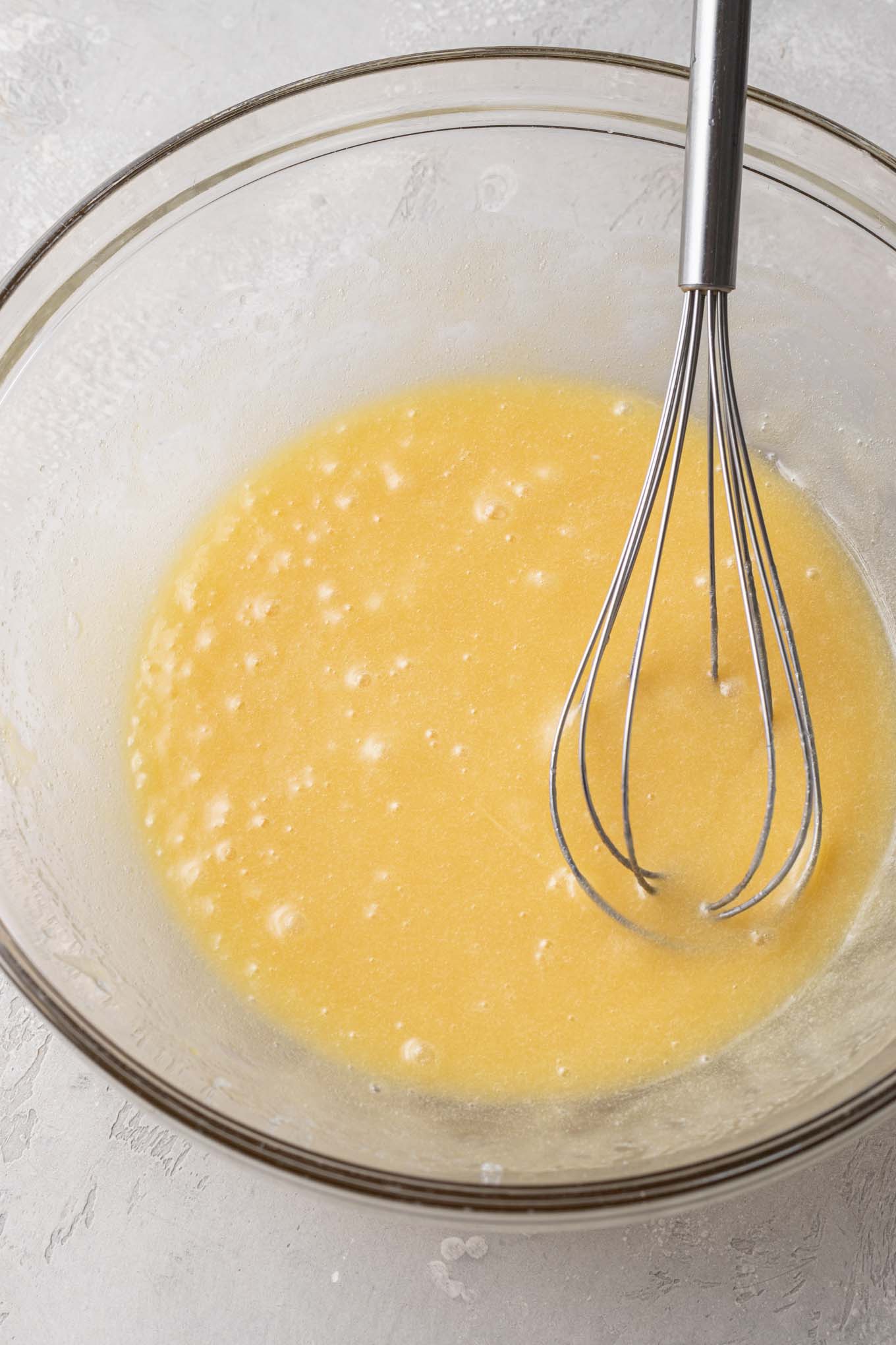 An overhead view of melted butter, eggs, and sugar in a glass mixing bowl with a whisk. 