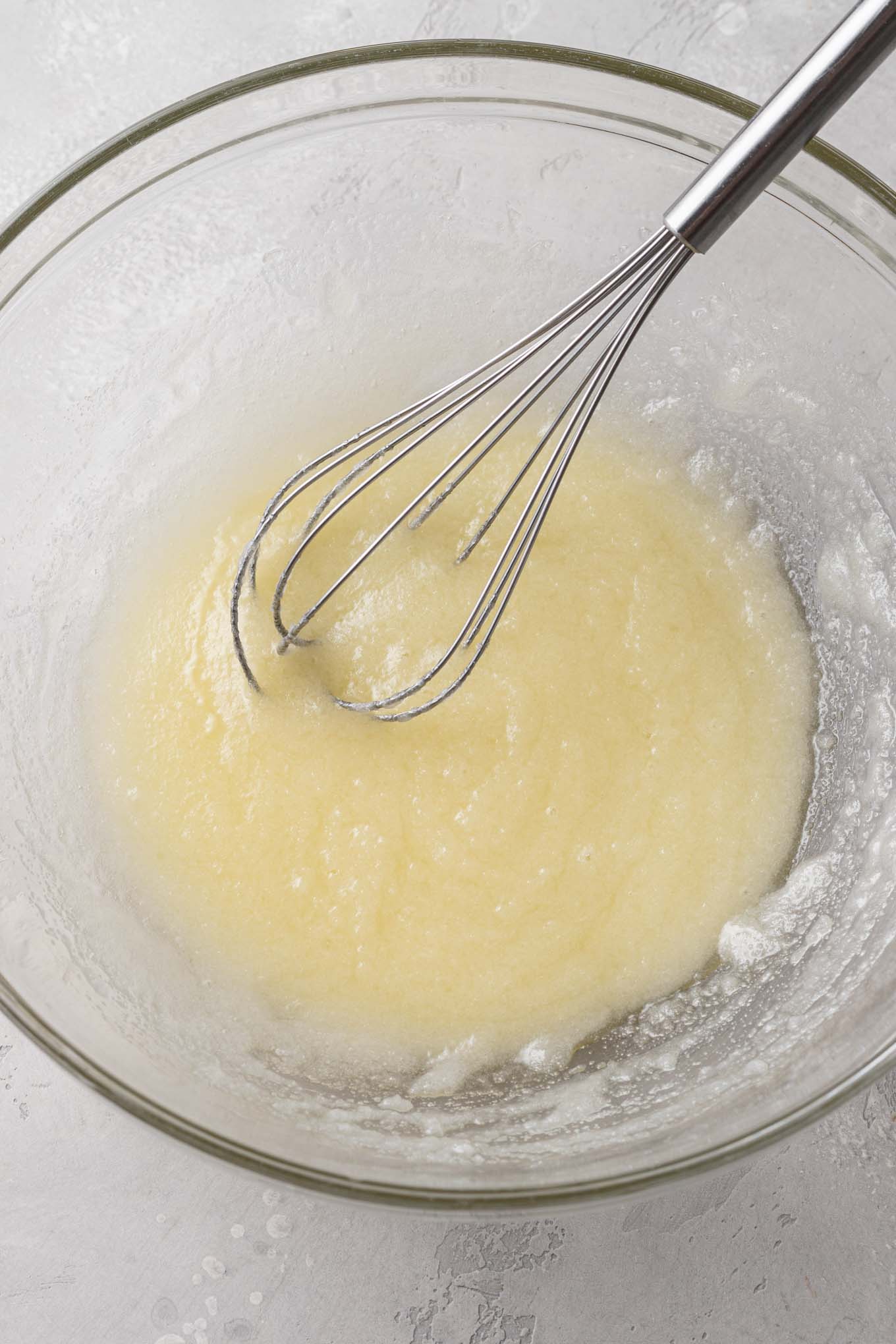 An overhead view of melted butter and sugar in a glass mixing bowl with a whisk. 