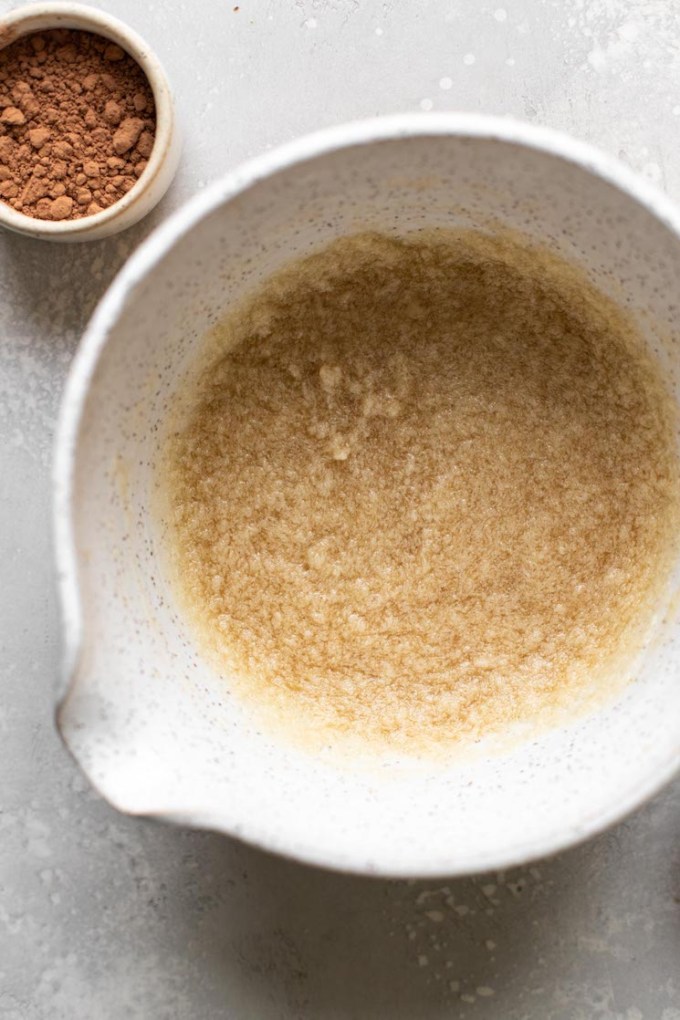 Overhead view of a mixing bowl with melted butter and sugar inside. A small dish of cocoa powder is on the side.