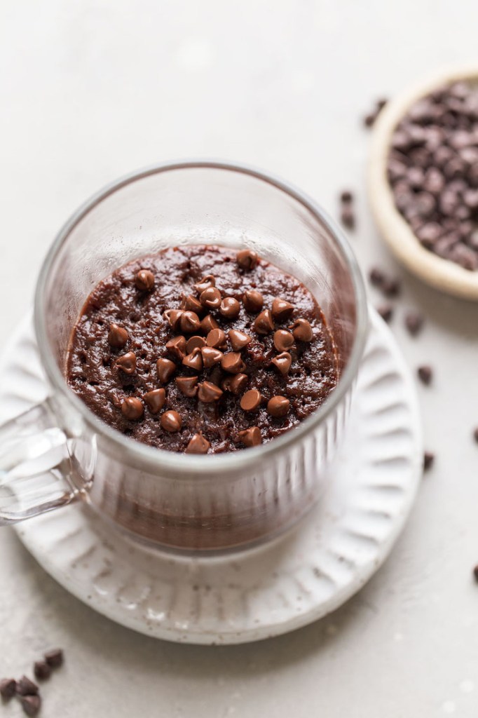 Overhead view of a microwave mug brownie in a glass mug. The mug sits on a white dessert plate, and a dish of mini chocolate chips rests on the side. 