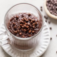 Overhead view of a microwave mug brownie in a glass mug. The mug sits on a white dessert plate, and a dish of mini chocolate chips rests on the side.