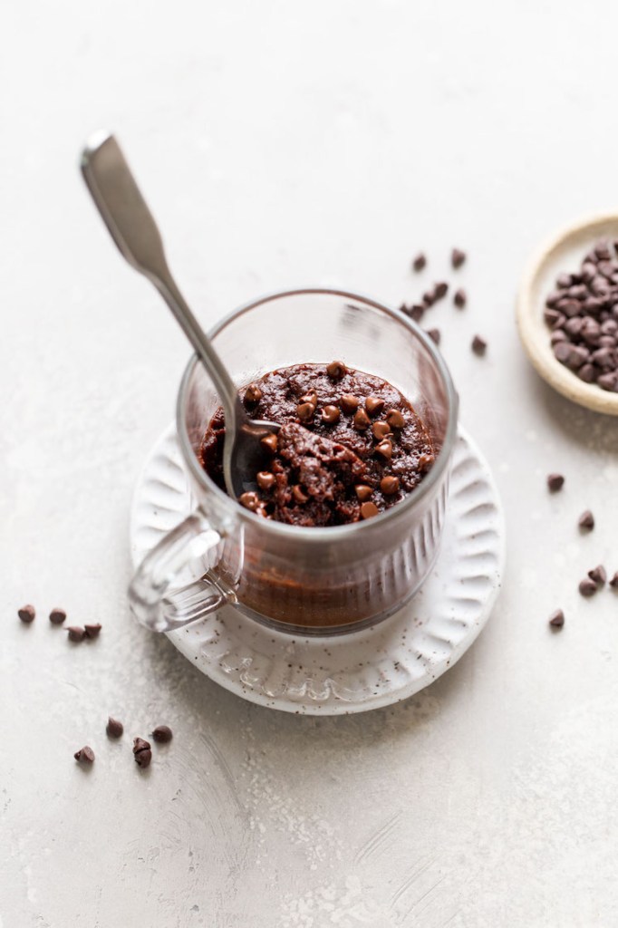 A gooey brownie in a mug, with a spoon sticking out. The mug is on a white dessert plate. Mini chocolate chips are scattered around, and they're also in a dish on the side. 