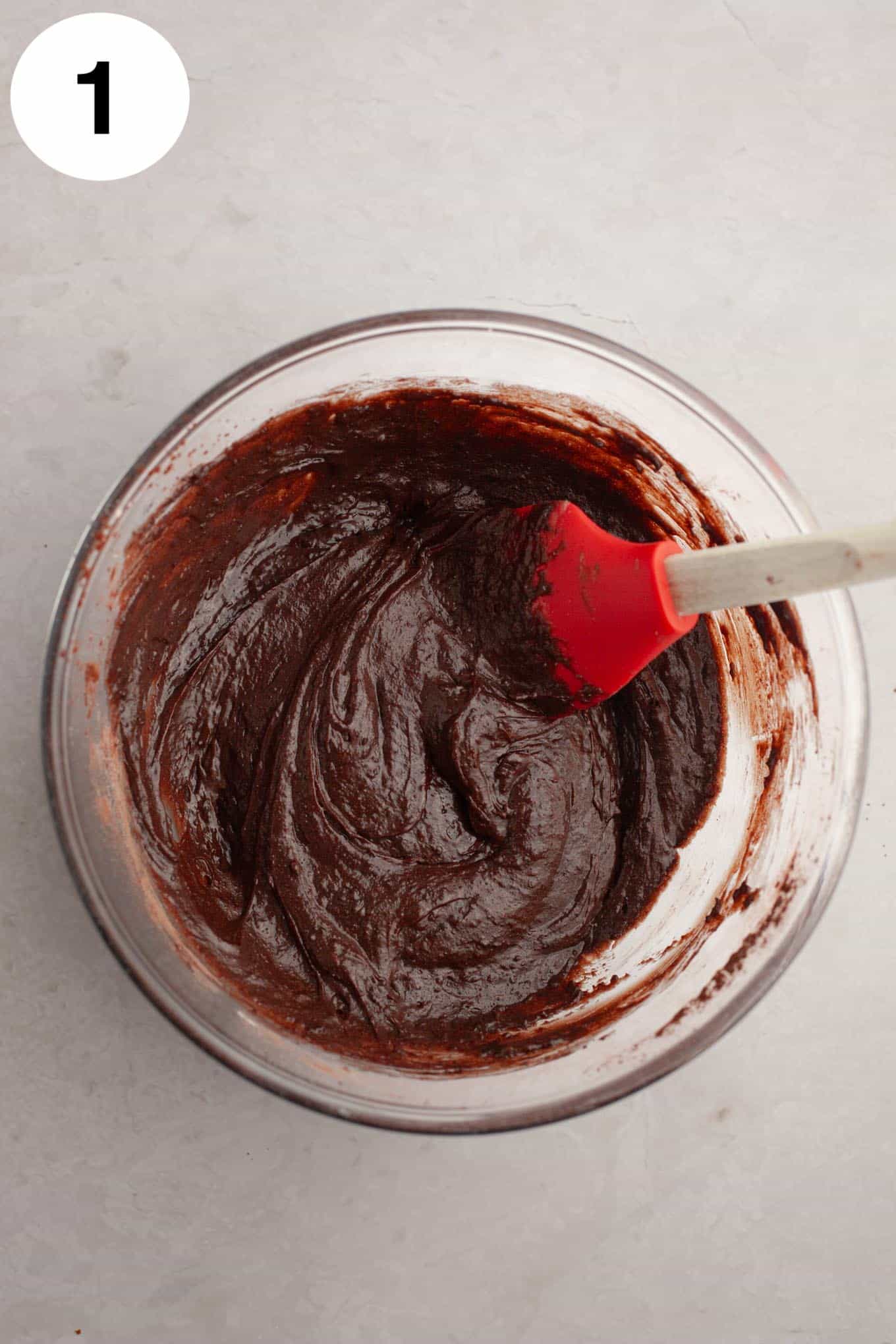 An overhead view of raw brownie batter in a glass mixing bowl, with a rubber spatula. 