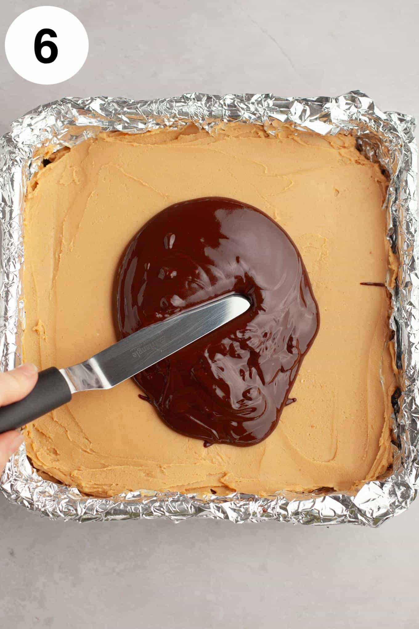 An overhead view of melted chocolate being spread over a layer of peanut butter fudge. 