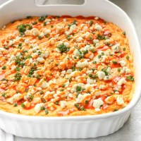 A white baking dish filled with creamy buffalo chicken dip topped with blue cheese, chives, and buffalo sauce.