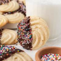 A butter cookie that has been dipped in chocolate sitting up against a glass of milk. More cookies and a bowl of sprinkles are around it.