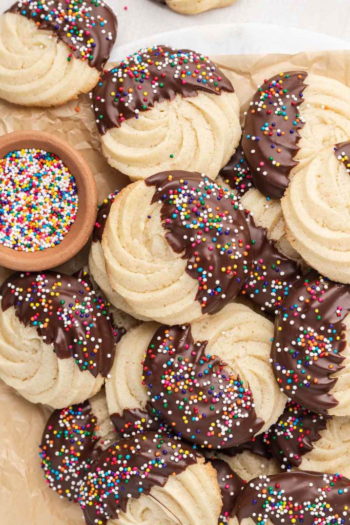 An overhead view of a pile of chocolate-dipped butter cookies with a small dish of sprinkles. 