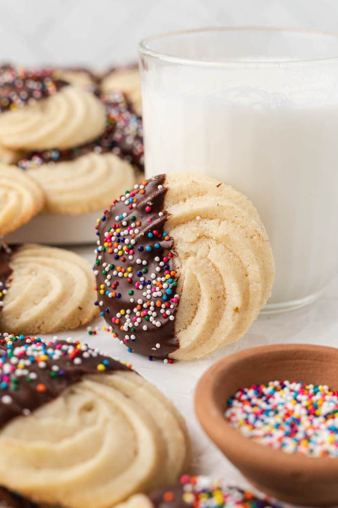 A chocolate-dipped butter cookie leaning against a glass of milk. More butter cookies surround them. 