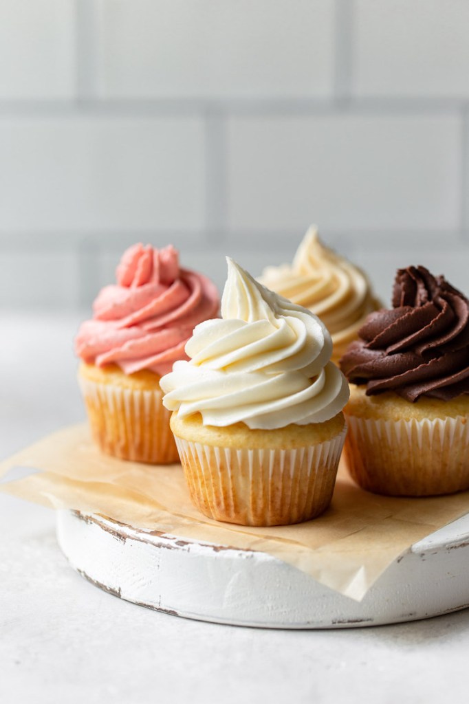 Four cupcakes on a rustic round piece of wood each topped with a different flavor of buttercream frosting.