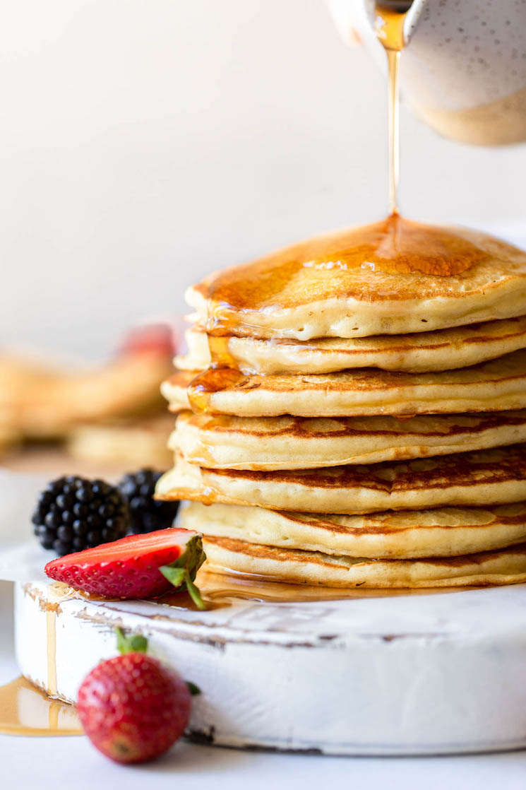 Maple syrup being poured onto a stack of pancakes on an antique white board with strawberries and blackberries surrounding it.