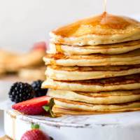 Maple syrup being poured onto a stack of pancakes on an antique white board with strawberries and blackberries surrounding it.