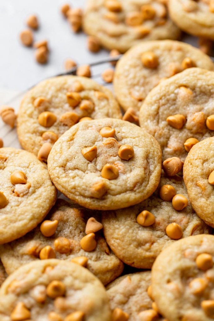 A close up view of butterscotch chip cookies piled on a wire cooling rack. Additional butterscotch chips are scattered in the background. 