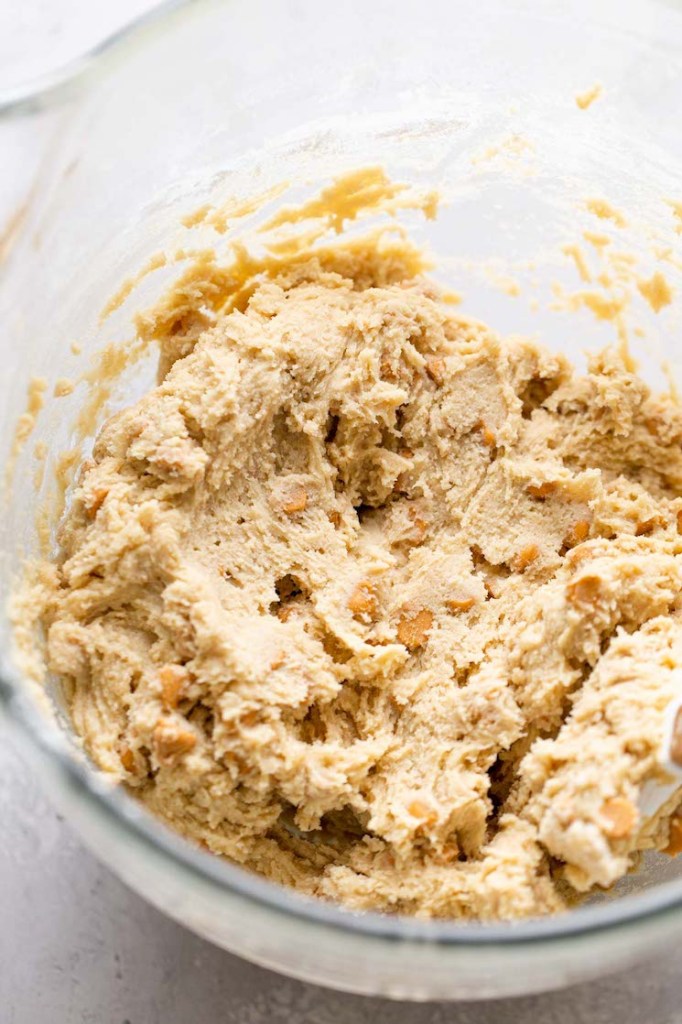 A glass mixing bowl of cookie dough with butterscotch chips in it. 