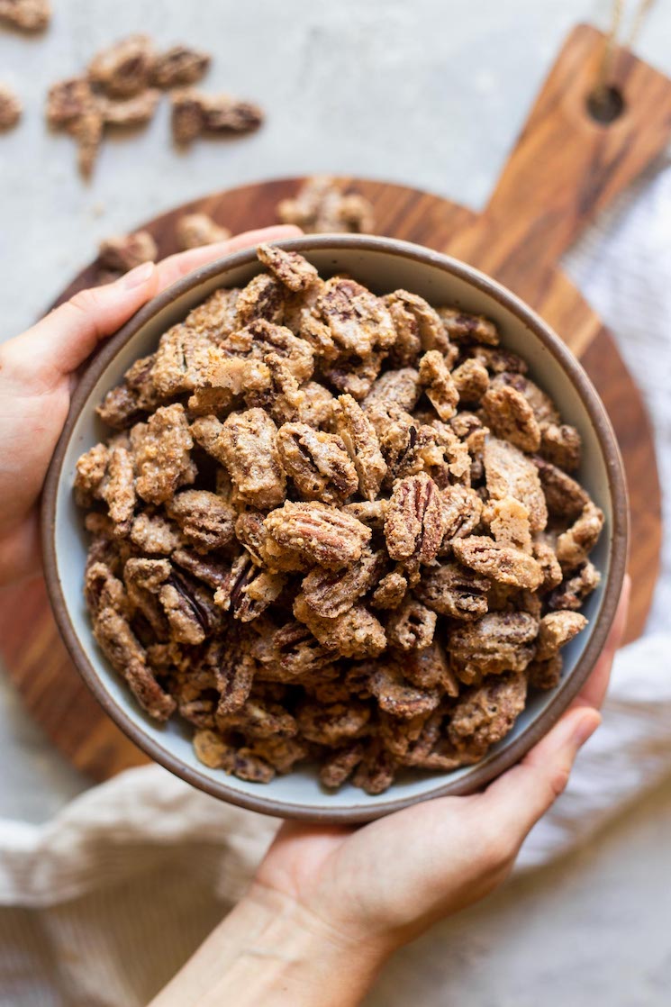 A bowl of candied pecans being held in the air over a round wooden tray and striped linen.