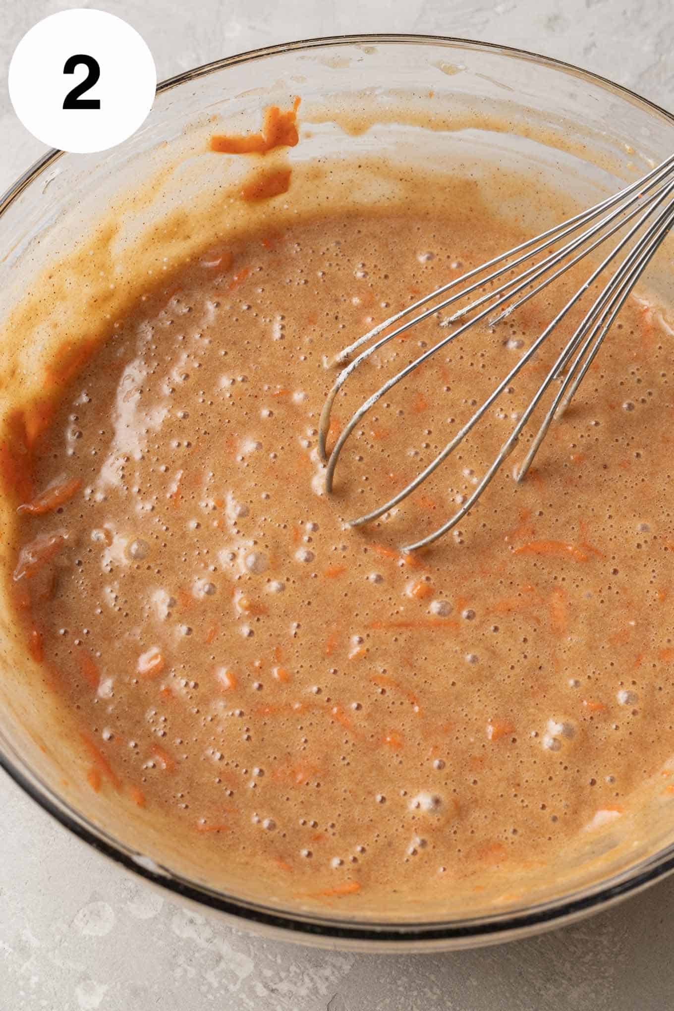 An overhead view of carrot cake batter in a glass mixing bowl with a whisk. 