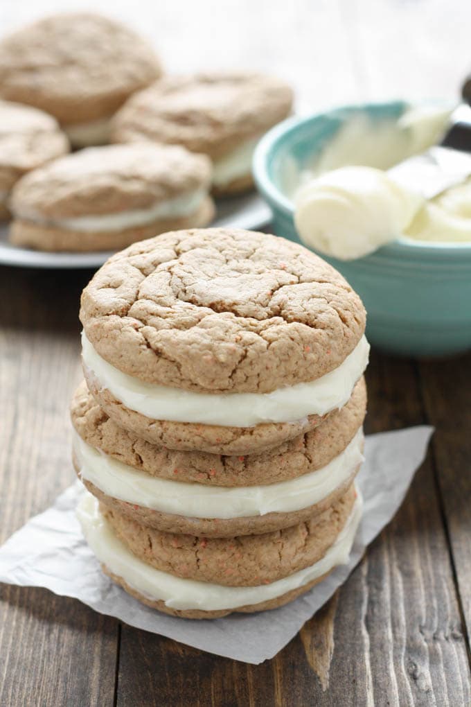 A stack of three carrot cake sandwich cookies on a square of parchment paper. A bowl of frosting and additional sandwich cookies rest in the background. 