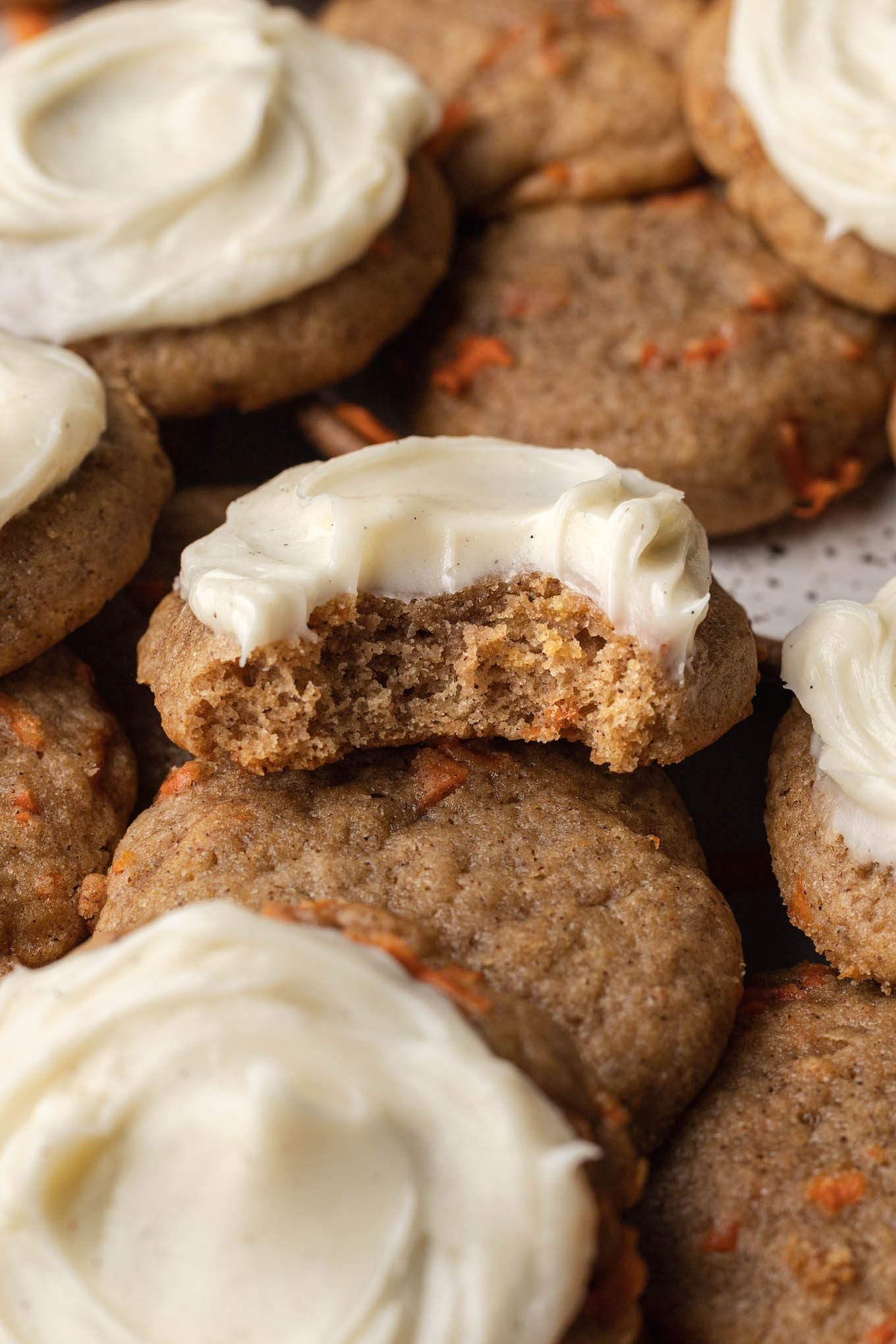 A close-up view of a frosted carrot cake cookie with a bite missing. 