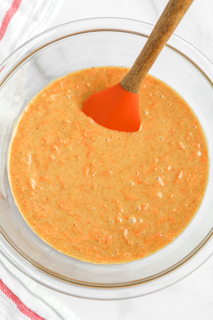 A glass bowl filled with carrot cake cupcake batter and a rubber spatula on the side.