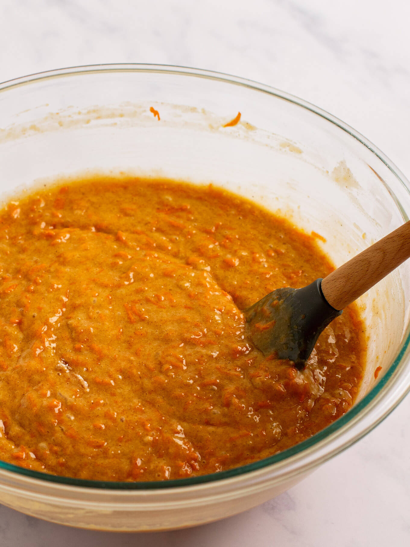 Carrot cake batter in a glass mixing bowl. A rubber spatula rests on the side of the bowl.