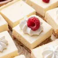 Several sliced cheesecake bars on a piece of parchment paper. Some of the bars are topped with whipped cream and a raspberry.