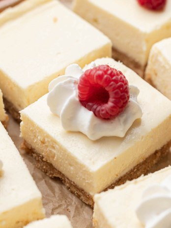 Several sliced cheesecake bars on a piece of parchment paper. Some of the bars are topped with whipped cream and a raspberry.