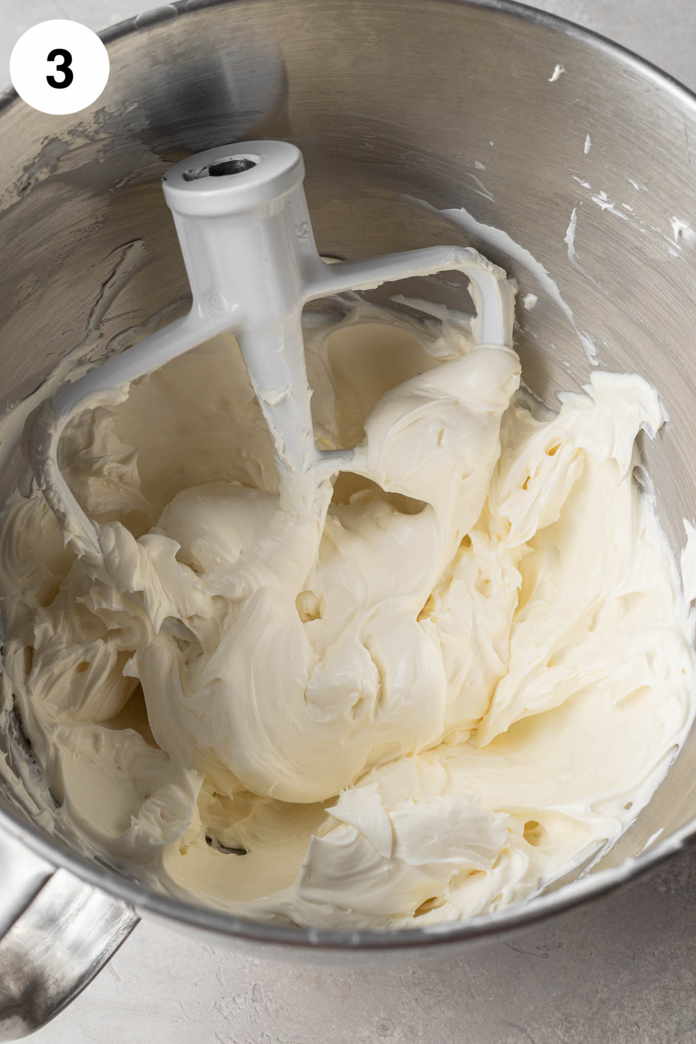 An overhead view of beaten cream cheese, sour cream, sugar, and vanilla in the bowl of a stand mixer, with a paddle attachment. 