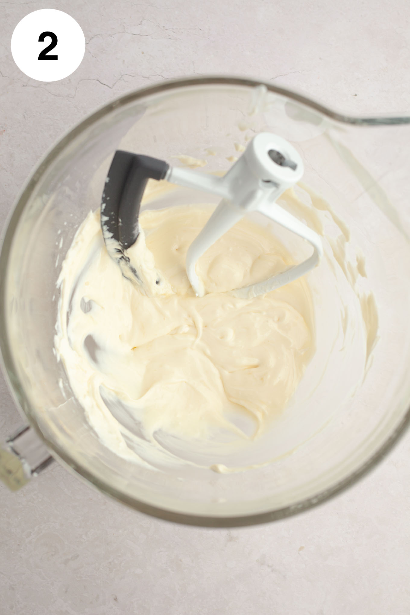 An overhead view of cream cheese beaten until smooth in a mixing bowl, with a paddle attachment. 