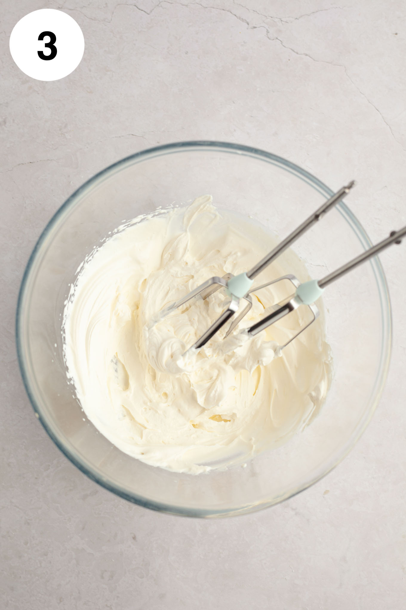 An overhead view of whipped cream in a glass mixing bowl, with two electric beaters. 