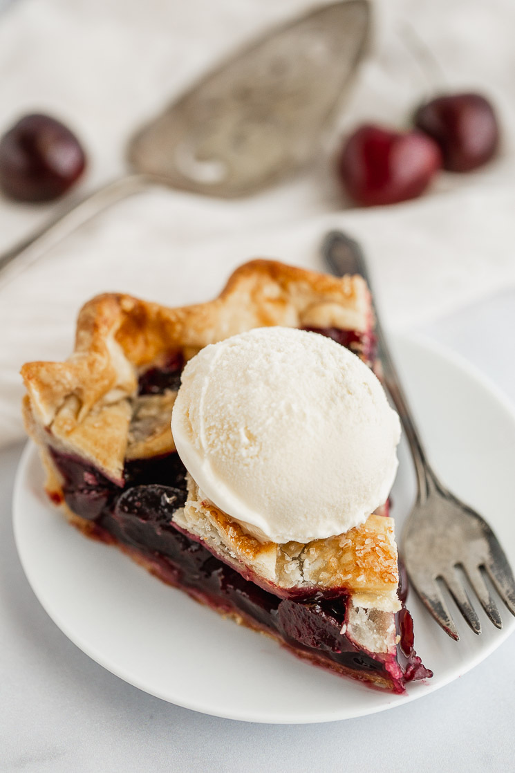 A slice of cherry pie with ice cream on top on a white plate.