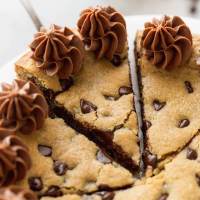 A cookie caked sitting on a white cake stand with a sliced being taken out of it.