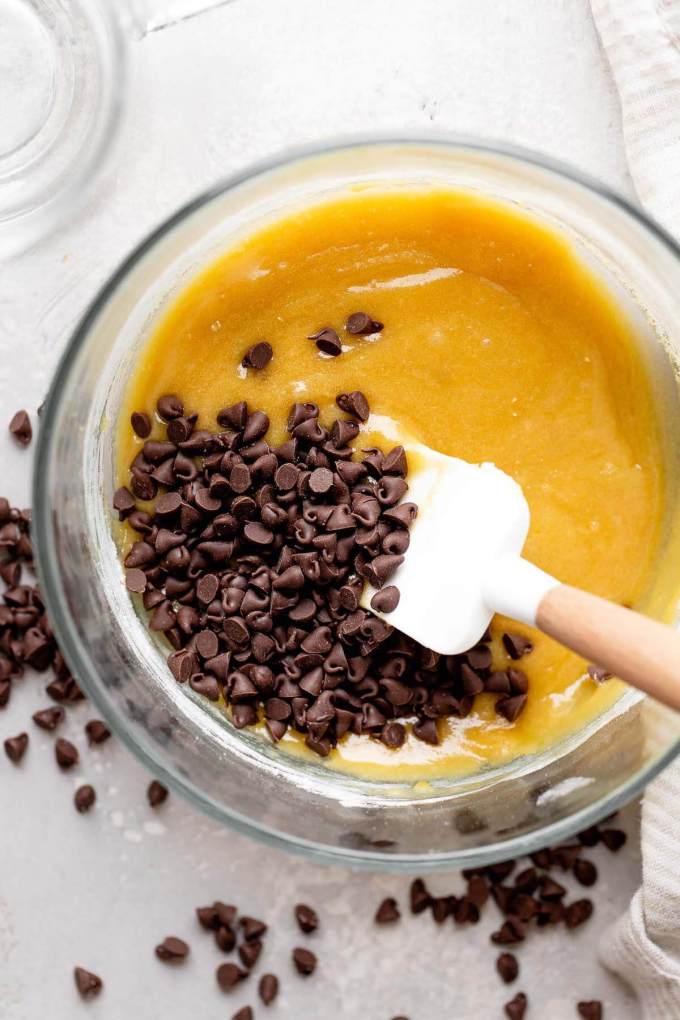 An overhead view of mini chocolate chips being stirred into raw cookie dough with a rubber spatula.