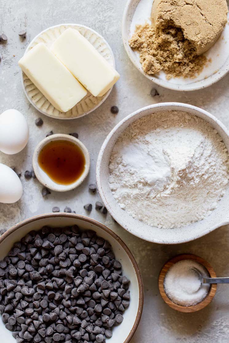 The ingredients needed to make chocolate chip cookies in bowls on top of a rustic gray surface.