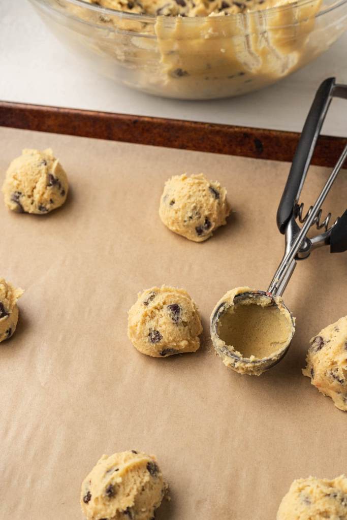 Chocolate chip cookie dough balls on a parchment-lined baking sheet, with a used cookie scoop. 