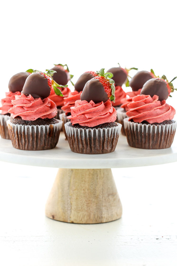 Chocolate cupcakes topped with strawberry buttercream frosting and chocolate covered strawberries on top of a white marble cake stand.