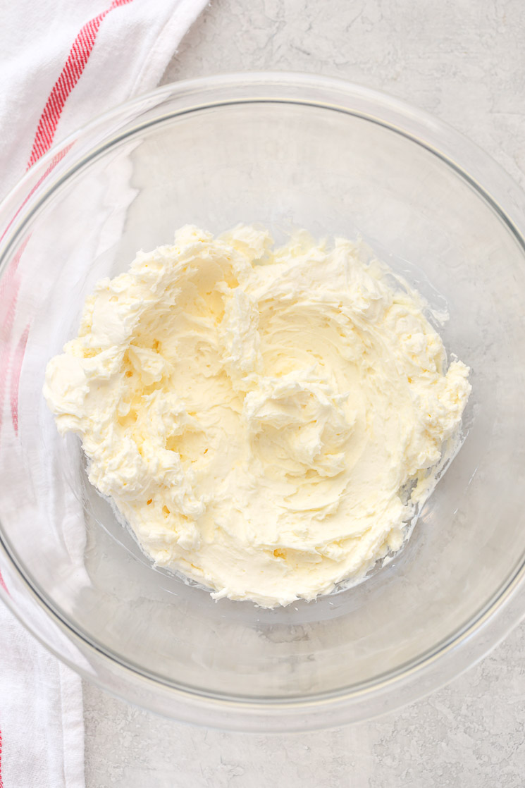 A clear glass mixing bowl holding butter and cream cheese combined together with a red and white napkin in the background. 