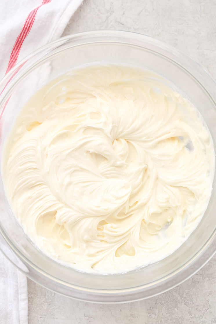 A clear glass mixing bowl holding cream cheese frosting ingredients with a white and red napkin in the background. 