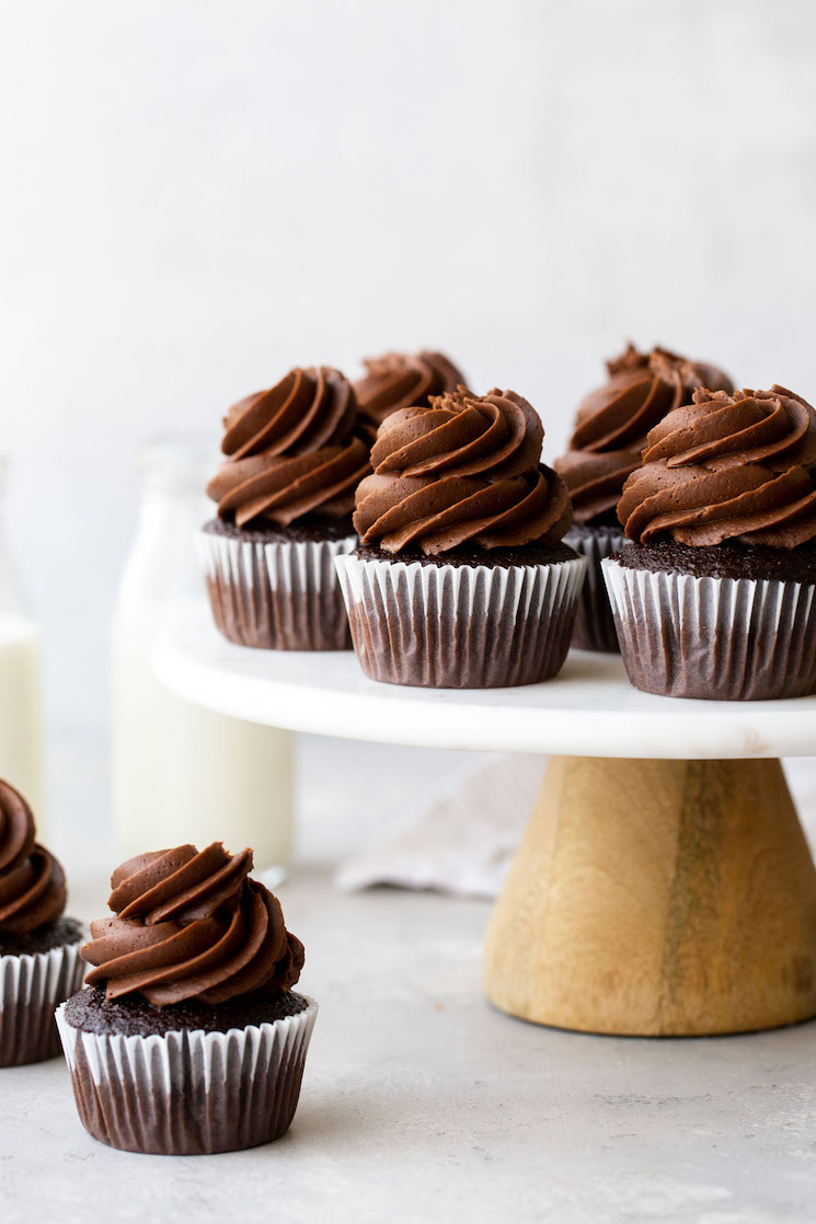 A marble cake stand holding chocolate cupcakes topped with chocolate frosting with jugs of milk in the background.