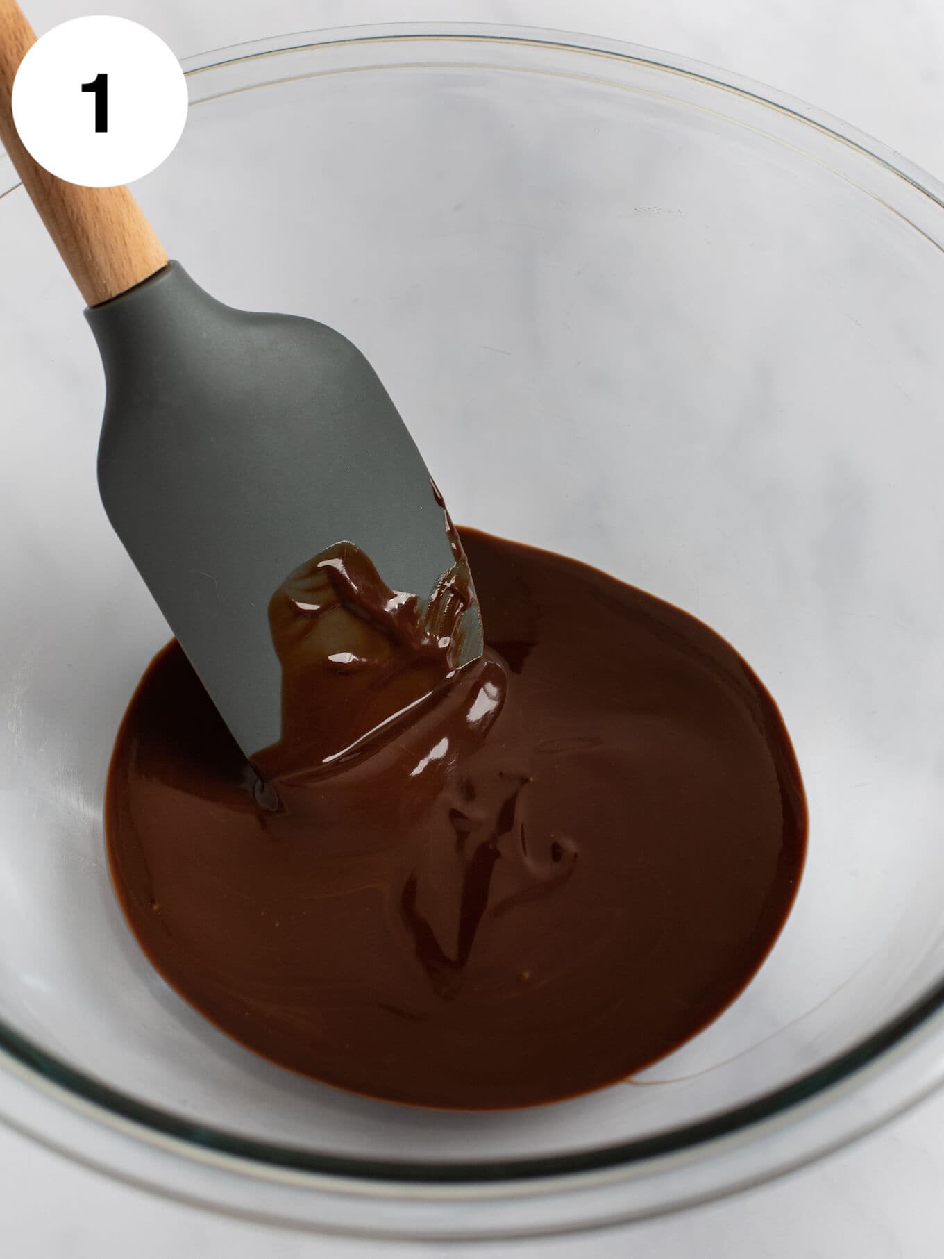 Melted chocolate and butter mixed together in a glass mixing bowl with a rubber spatula.