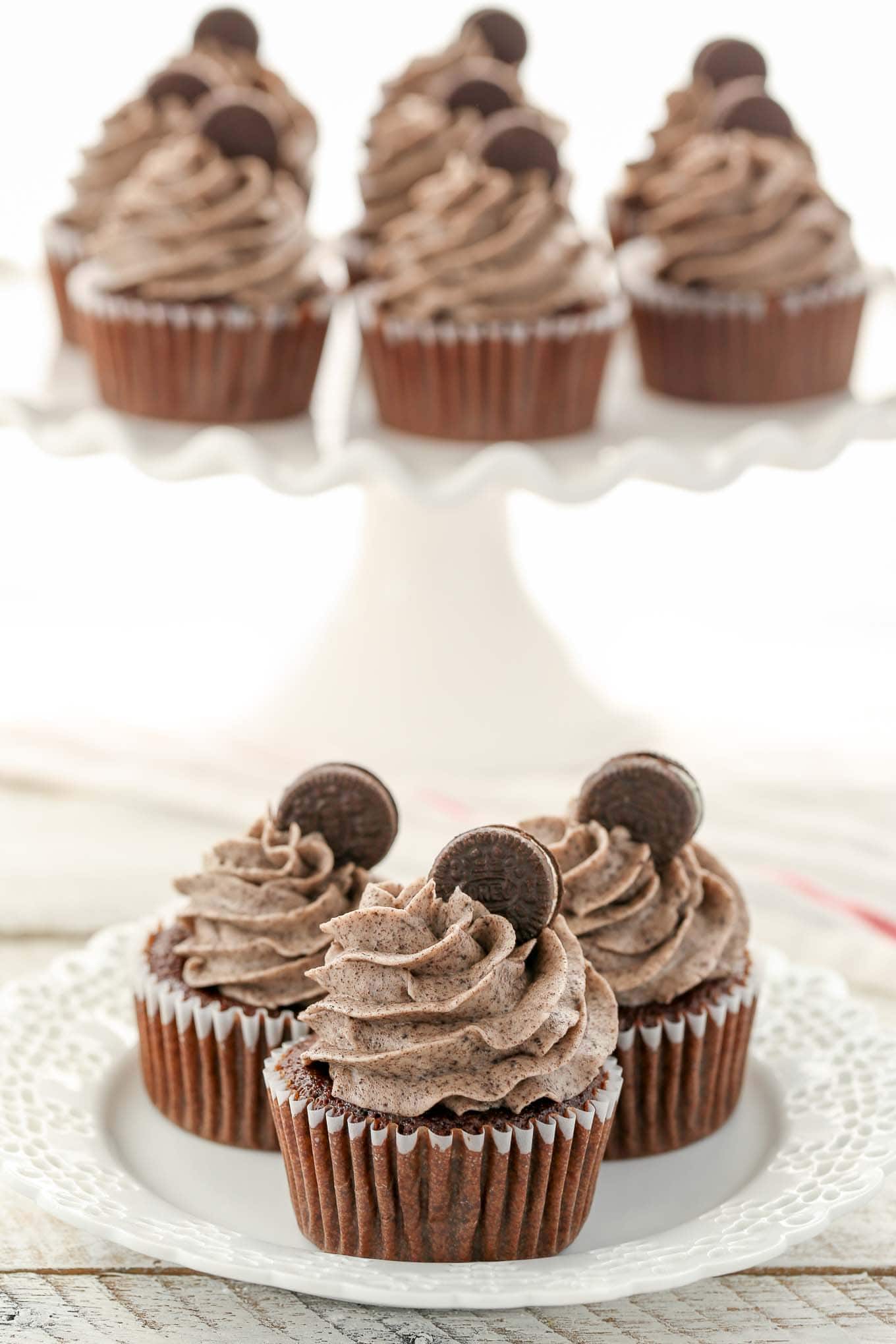 Three oreo cupcakes on a white plate. More cupcakes rest on a cake stand in the background. 