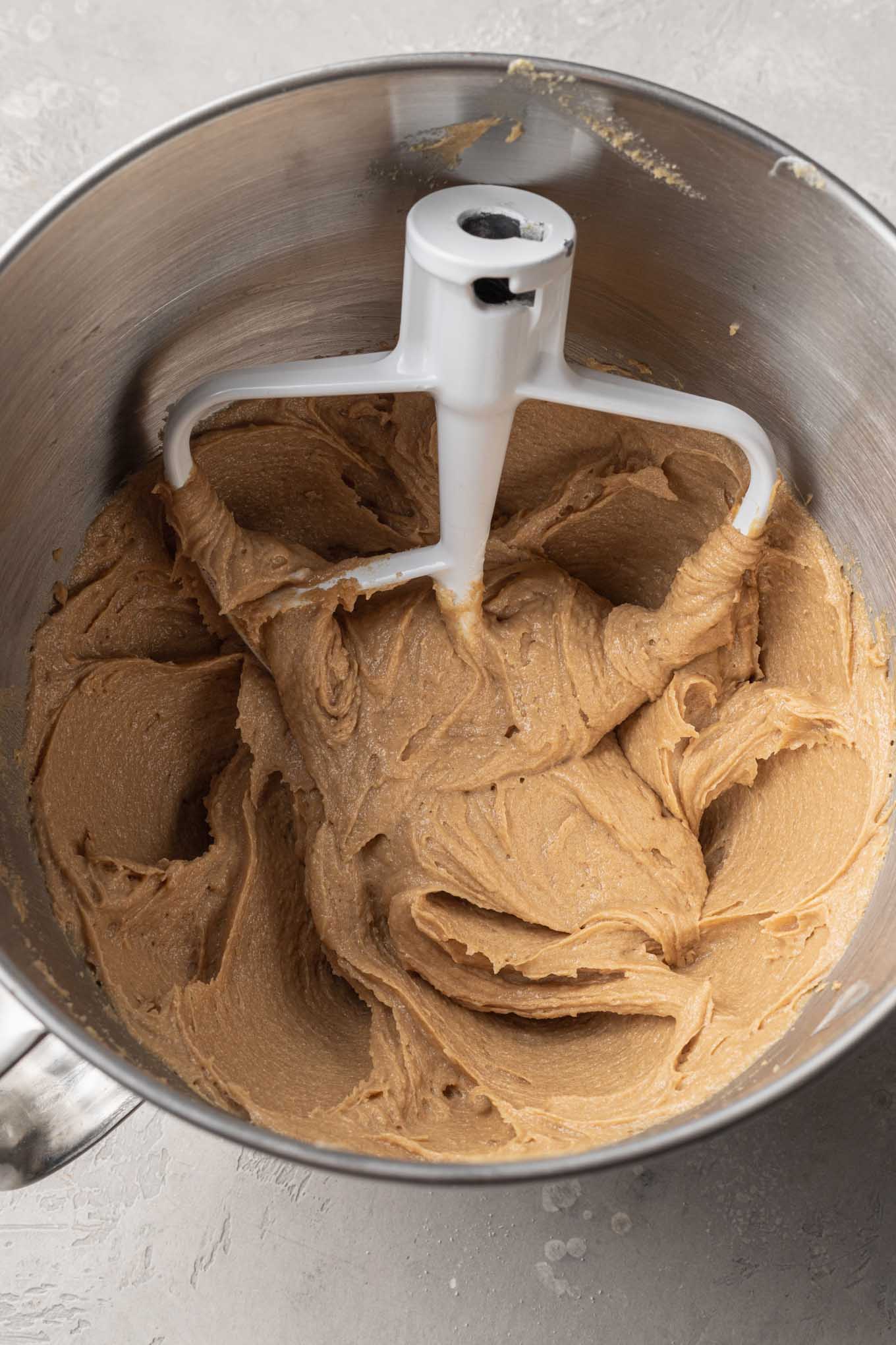 An overhead view of a peanut butter mixture in the bowl of a stand mixer, with a paddle attachment. 