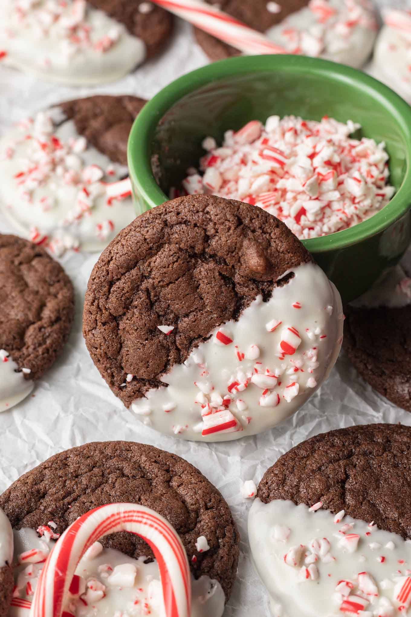 A peppermint chocolate cookie resting against a bowl of candy cane pieces, surrounded by additional cookies. 