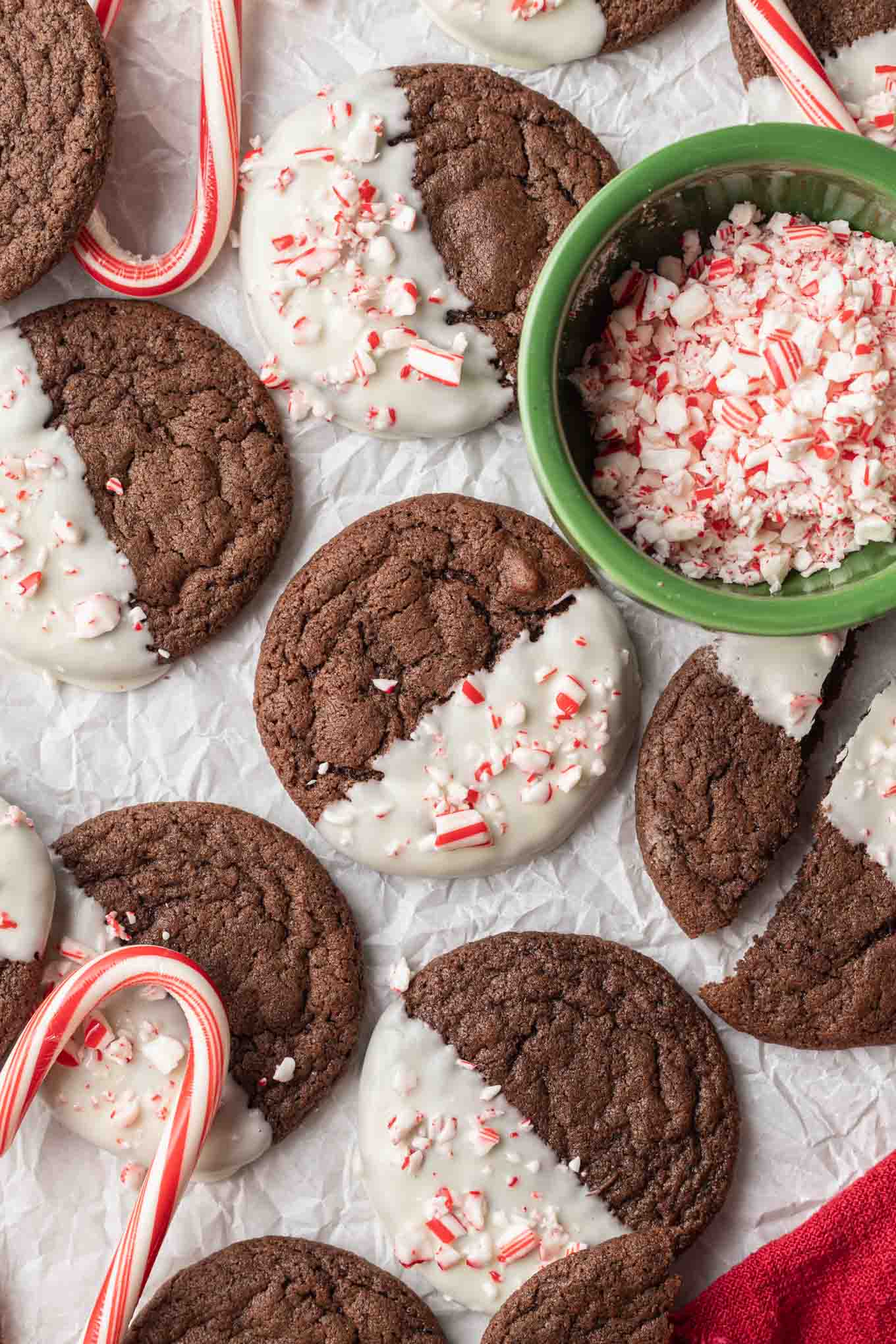 An overhead view of white chocolate-dipped chocolate peppermint cookies, with a dish of crushed candy canes. 