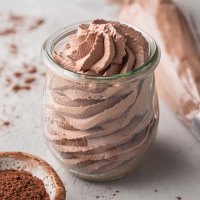 A small jar filled with chocolate whipped cream. A piping bag filled with whipped cream and a small bowl of cocoa powder rest nearby.